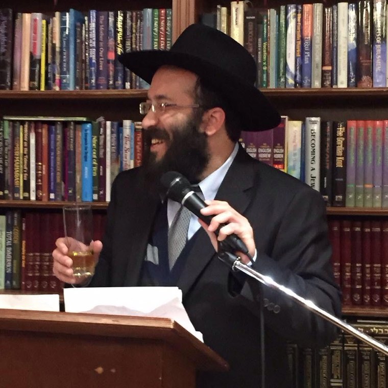 Rabbi Nachmi Deitsch speaking, holding a small glass, before shelves of English Judaica